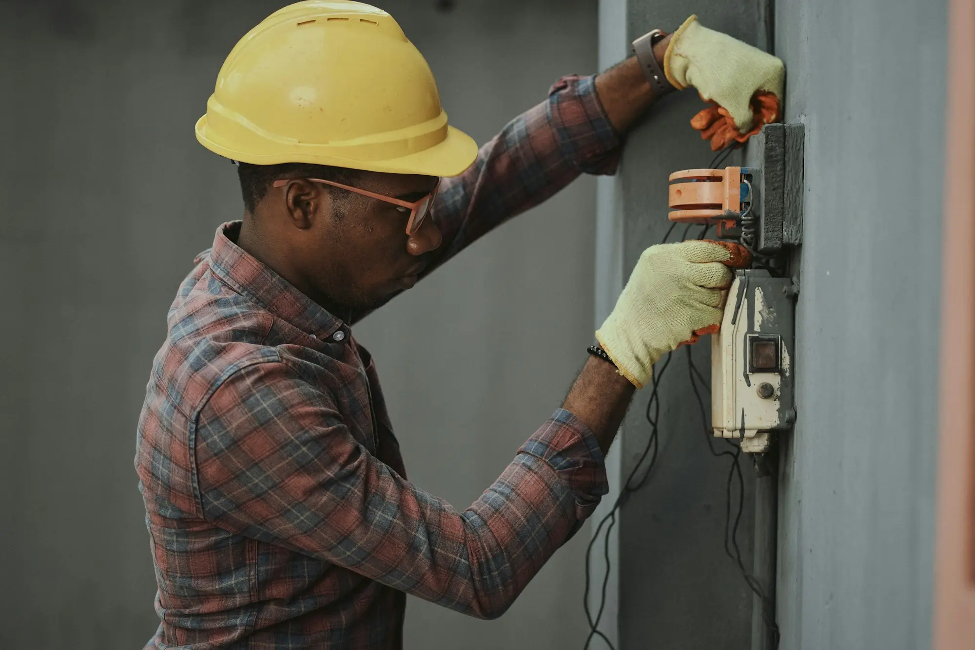 Electrician in a yellow hard hat and plaid shirt holding equipment on a job site